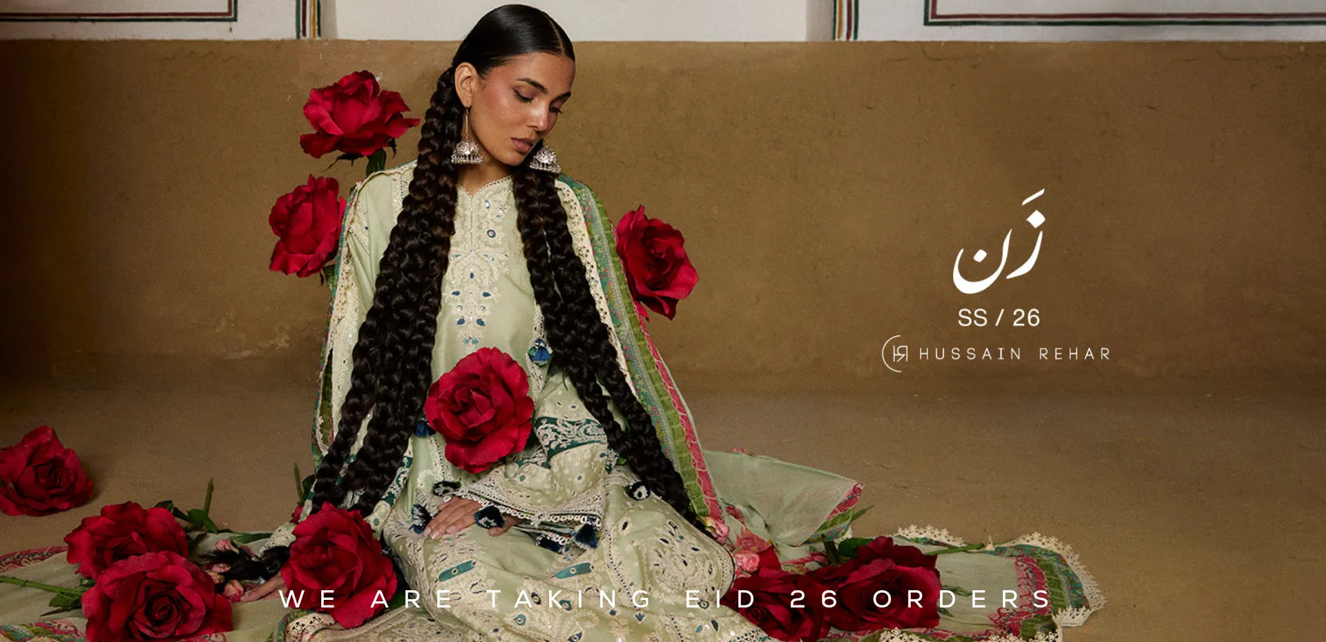Woman in traditional Pakistani outfit with red flowers, sitting on a brown couch with 'Hussain Behar' branding Eid Dresses UK.1621243260e1af0c20-1